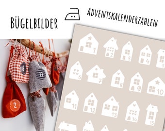 May include: A set of 15 white house-shaped iron-on transfers for an advent calendar. The transfers are numbered 1 through 15 and feature different designs, including stars, hearts, and windows. The transfers are on a beige background.