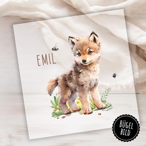 Op de afbeelding: Een aquarel-illustratie van een schattig wolfje met bruine vacht en een witte borst. De wolf staat in een veld met groen gras en gele bloemen. Er vliegen twee bijen rond de wolf. De tekst "EMIL" staat in bruine letters bovenaan de afbeelding. De woorden "BÜGEL BILD" staan in een cirkel onderaan de afbeelding.