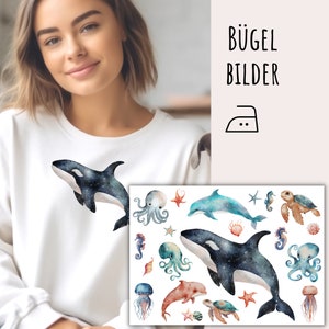 Op de afbeelding: Witte sweatshirt met een aquarel orka walvis ontwerp met een sterrenhemel patroon. Inclusief een strijktransfervel met diverse zeedieren, waaronder dolfijnen, octopussen en zeeschildpadden. De tekst "BÜGEL BILDER" is zichtbaar.