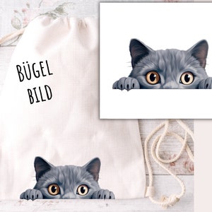 Può includere: Una borsa a cordoncino bianca con la scritta "BÜGEL BILD" stampata sul davanti. Un gatto grigio con gli occhi gialli sta guardando da sopra la borsa.