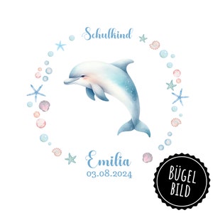 May include: A watercolor illustration of a blue dolphin jumping out of the water. The dolphin is surrounded by a circle of pink and blue bubbles, seashells, and starfish. The text "Schulkind" is at the top of the circle, and the text "Emilia 03.08.2024" is at the bottom. The text "BÜGEL BILD" is in a black circle at the bottom right corner.