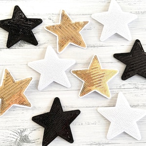 Könnte beinhalten: Sortiment von acht sternförmigen Bügelbildern in Schwarz, Weiß und Gold. Die Patches sind aus Pailletten gefertigt und haben eine glänzende Oberfläche.