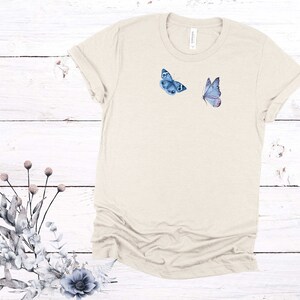 May include: A light beige t-shirt with two blue butterflies printed on the front. The butterflies are in a watercolor style.
