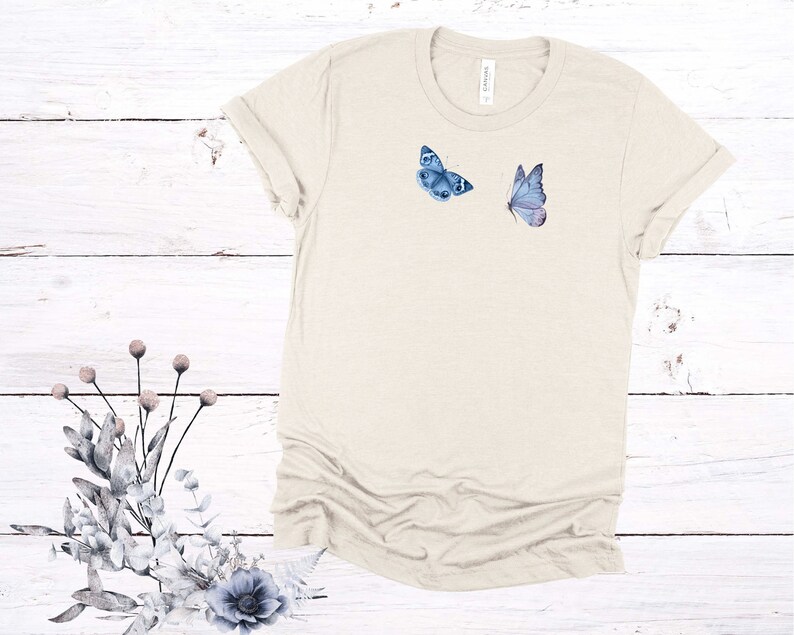 May include: A light beige t-shirt with two blue butterflies printed on the front. The butterflies are in a watercolor style.