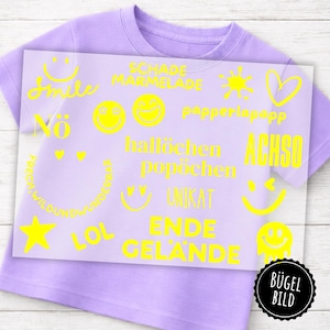 Può includere: Una maglietta lilla con una sovrapposizione grafica gialla. La sovrapposizione presenta varie faccine sorridenti, stelle, cuori e testo in tedesco, tra cui "Smile", "LOL" e "Ende Gelände". Un logo circolare in bianco e nero si trova nell'angolo in basso a destra.