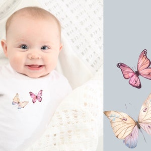 May include: A white onesie with a pink and white butterfly design. The onesie is worn by a baby with a happy expression.