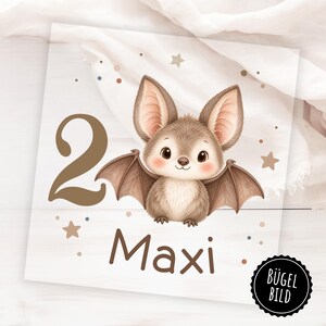 May include: A square iron-on transfer featuring a brown cartoon bat, the number 2, and the name Maxi in brown text. The design includes small brown stars and dots, with a black circular badge in the corner containing the text BÜGEL BILD.
