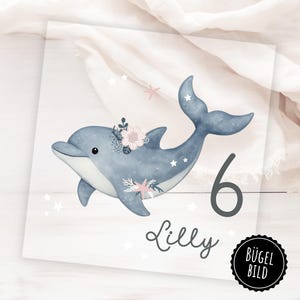 Op de afbeelding: Waterverfillustratie van een blauwe dolfijn versierd met bloemen en zeesterren. Het cijfer 6 en de naam "Lilly" staan onder de dolfijn geschreven. Witte sterren zijn verspreid rond de dolfijn. Een zwart-witte cirkel met de tekst "BÜGEL BILD" staat rechtsonder.