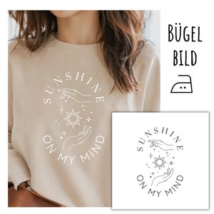 May include: Beige sweatshirt featuring the text "SUNSHINE ON MY MIND" in a circular design. The design includes hands, a sun, and stars. The image also includes a smaller version of the design and an iron symbol.