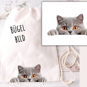 Può includere: Una borsa a cordoncino bianca con la scritta "BÜGEL BILD" stampata sul davanti. Un gatto grigio con gli occhi arancioni spunta da dietro la borsa.