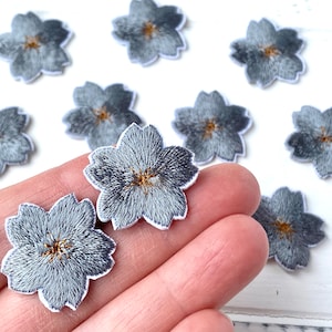 Könnte beinhalten: Zehn graue Filzblumen-Patches mit goldenen Zentren. Die Blumen sind auf einer weißen Oberfläche angeordnet und eine Hand hält zwei der Patches.