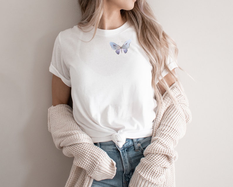 May include: A white t-shirt with a watercolor butterfly design on the front. The butterfly is a light blue color with hints of purple and gray.