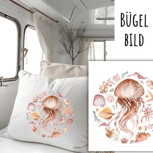 May include: A white pillow featuring a watercolour design of a jellyfish and seashells in shades of pink, orange, and brown. The design is in a circular shape and the text "BÜGEL BILD" is in the top right corner of the image.