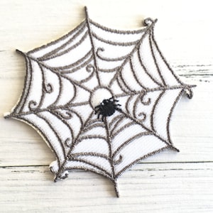 Può includere: Una toppa bianca ricamata a forma di ragnatela con un ragno nero al centro.