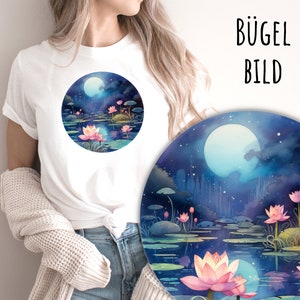 Op de afbeelding: Een wit T-shirt met een rond ontwerp van een maanverlichte vijver met roze lotusbloemen. Het ontwerp is omgeven door een donkerblauwe rand.