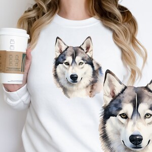 May include: White sweatshirt with a watercolor illustration of two husky dogs. The dogs are facing forward with their eyes open and their tongues out. The sweatshirt is a light white color and the dogs are a light gray and white color.