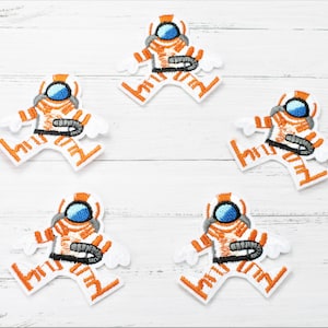 Könnte beinhalten: Fünf orangefarbene und weiße gestickte Bügelbilder von Astronauten in Raumanzügen. Die Patches befinden sich auf einem weißen Holzgrund.