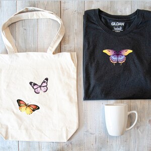May include: A white tote bag with three butterfly designs. The butterflies are pink, orange, and purple. The bag is next to a black Gildan t-shirt with a purple butterfly design. A white mug is also in the image.