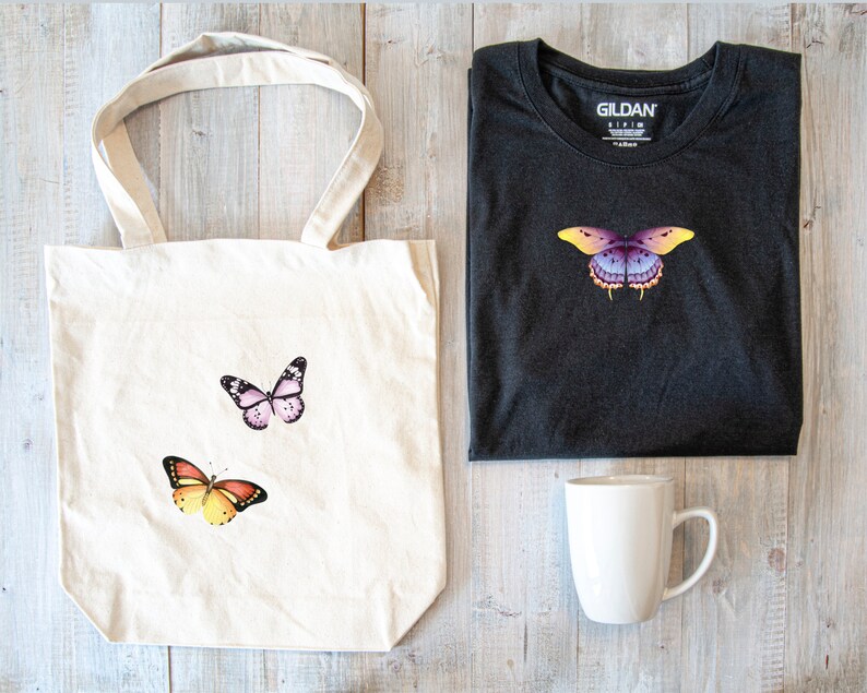 May include: A white tote bag with three butterfly designs. The butterflies are pink, orange, and purple. The bag is next to a black Gildan t-shirt with a purple butterfly design. A white mug is also in the image.