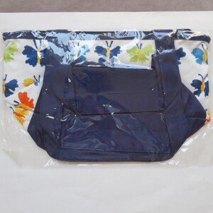 May include: A tote bag with a white background and a butterfly pattern in blue, orange, and yellow. The bag has a navy blue interior and handles. The bag is sealed in clear plastic.