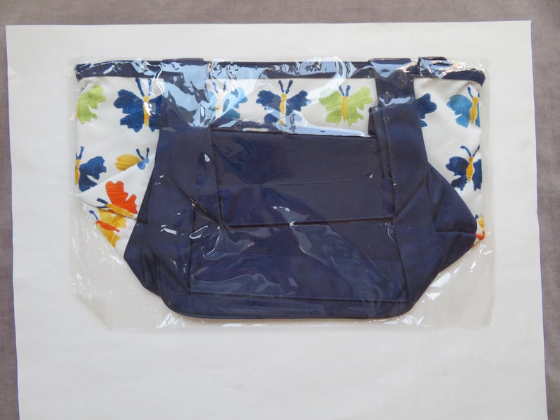 May include: A tote bag with a white background and a butterfly pattern in blue, orange, and yellow. The bag has a navy blue interior and handles. The bag is sealed in clear plastic.
