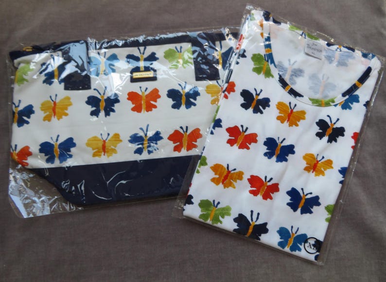 May include: A white tote bag and a white t-shirt, both with a repeating pattern of colorful butterflies in orange, yellow, blue, and green. The tote bag has a navy blue trim.