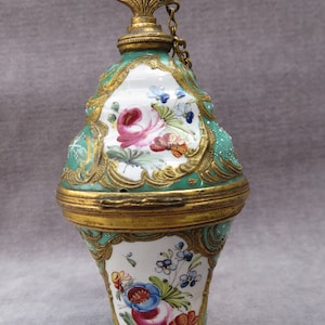 May include: A green and gold porcelain perfume bottle with a floral design and a gold chain. The bottle is decorated with gold accents and has a floral design on the bottom.