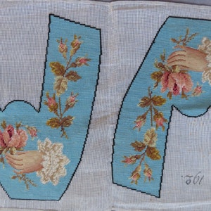 May include: Two blue canvas panels with embroidered letters "W" and "A" in black. Each letter is decorated with a hand holding a pink rose and white flowers, surrounded by green leaves and brown stems. The panels are signed "261" in the bottom right corner.