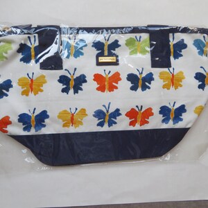 May include: A white tote bag with a repeating pattern of colorful butterflies in blue, yellow, orange, and green. The bag has a navy blue bottom and handles, and is packaged in clear plastic.
