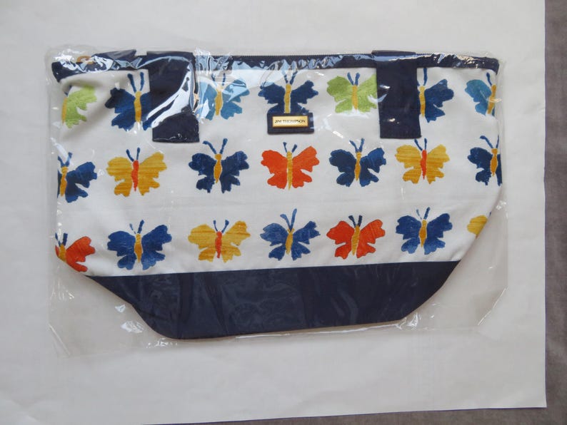 May include: A white tote bag with a repeating pattern of colorful butterflies in blue, yellow, orange, and green. The bag has a navy blue bottom and handles, and is packaged in clear plastic.