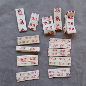 May include: A collection of off-white fabric clothing labels featuring various red embroidered monograms and initials including DL, BH, DD, BL, WR, WP, OG, UZ, OP, TP, RD, VJ, and HO. These rectangular woven tags are arranged in a scattered layout.