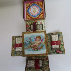 May include: A vintage needle case with a cherub design and the text "The Alexandra Needle Case". The case is made of cardboard and has a floral pattern. It has six compartments for needles, each labeled with a number from 6 to 8.