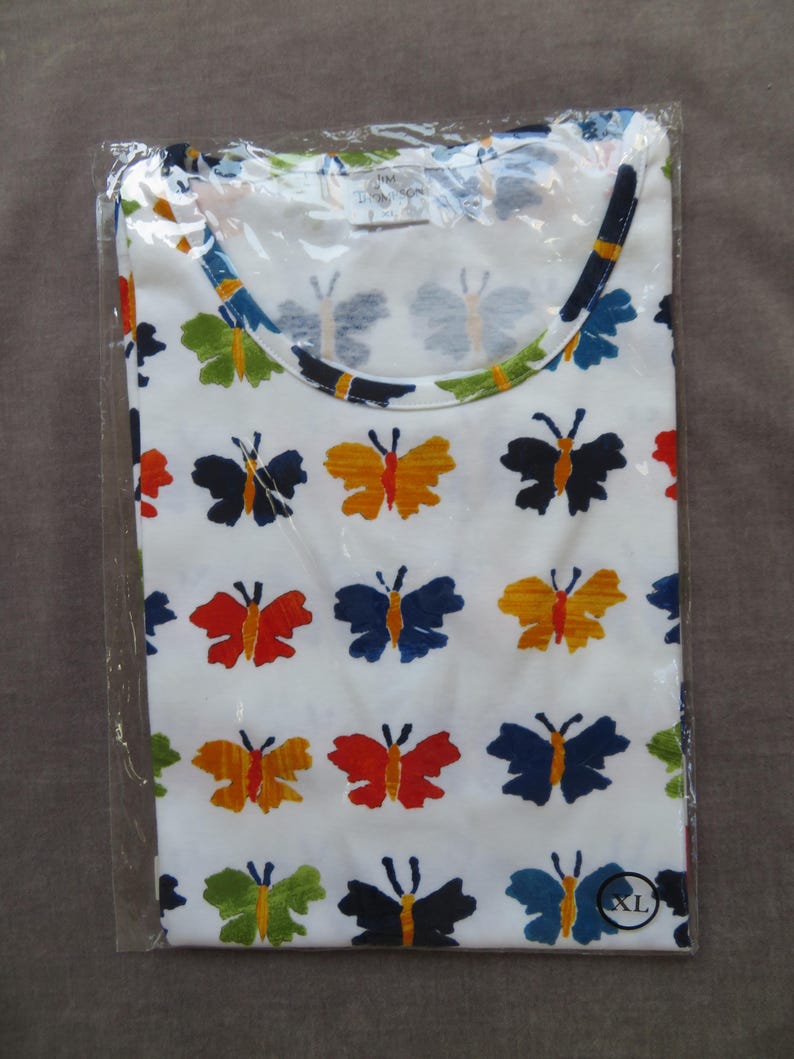 May include: White t-shirt with a colorful butterfly pattern. The butterflies are in various colors, including red, orange, blue, green, and black. The shirt is folded and sealed in a clear plastic bag. The shirt has a round neckline.