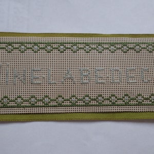 May include: A rectangular needlepoint canvas with the words "SINE LABE DEUS" stitched in light grey thread. The canvas is bordered with a green geometric pattern and rests on a green fabric.