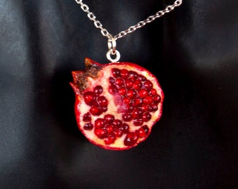 Könnte beinhalten: Eine silberfarbene Halskette mit einem Anhänger in Form eines halbierten Granatapfels. Der Anhänger zeigt einen realistischen Querschnitt der Frucht mit tiefroten Kernen und einer strukturierten, rotbraunen Schale am oberen Rand.