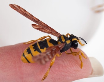 May include: A miniature wasp figurine featuring a yellow and black striped abdomen, a black thorax, and translucent brown wings. The insect model has thin, articulated legs and a detailed head with black eyes, shown in a small scale.