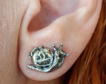 Könnte beinhalten: Ein silberfarbener Ohrstecker in Form einer Schnecke mit einem geschnitzten Kürbisgehäuse. Die Schnecke hat zwei kleine Fühler und einen strukturierten Körper, während das Gehäuse ein geschnitztes Kürbisgesicht mit Aussparungen zeigt.