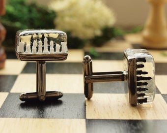 Könnte beinhalten: Ein Paar quadratische Manschettenknöpfe mit einem klaren Harzblock, der Miniatur-Schachfiguren in Weiß und Schwarz umschließt. Die Manschettenknöpfe haben eine metallisch silberfarbene Basis mit einem standardmäßigen Knebelverschluss.