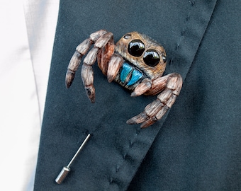Könnte beinhalten: Eine spinnenförmige Anstecknadel mit braun texturiertem Körper, großen schwarzen glänzenden Augen und leuchtend metallisch blauen Kieferklauen. Die Spinne hat acht segmentierte Beine und ist an einer silberfarbenen Metallnadel befestigt.