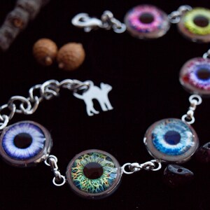 May include: A silver-toned bracelet featuring multiple circular charms with detailed eye designs in various colors, including blue, green, and pink. The bracelet has a chain link design and a charm with a cat silhouette. The background is black.