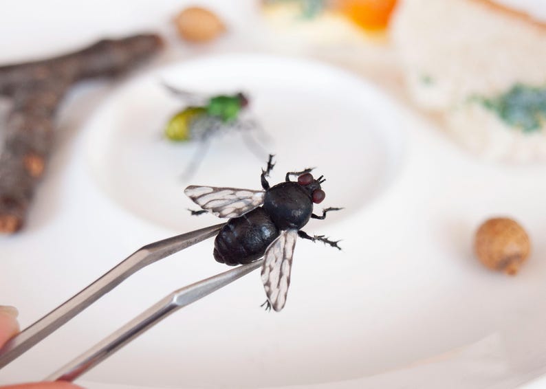 May include: A pair of silver metal tweezers holding a realistic black fly figurine with translucent white wings and dark red eyes. A second smaller green and yellow fly figurine is visible in the background on a white surface.