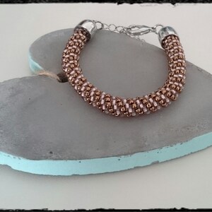 Könnte beinhalten: Ein Armband aus roségoldenen und weißen Perlen mit einem silbernen Verschluss, auf einer grauen herzförmigen Oberfläche präsentiert.