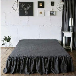 May include: A gray linen bed skirt with a ruffled edge. The bed skirt is on a bed with white walls and a gray curtain in the background.
