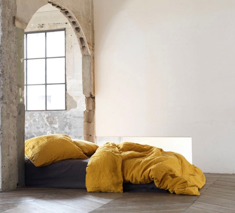 May include: A mustard yellow linen duvet cover and pillowcases on a bed with a gray linen sheet. The bed is in a room with a large window and exposed brick walls.