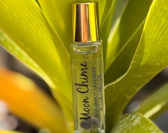 May include: A clear glass essential oil roller bottle with a gold cap, labeled "Moon Chime" with "Bergamot, Lavender & Coconut Oil" text. The bottle is set against a backdrop of vibrant green leaves, creating a natural and fresh aesthetic.