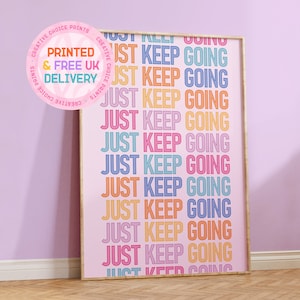Puede incluir: Un póster rosa con el texto "JUST KEEP GOING" repetido en colores arcoíris. El póster está enmarcado en un marco dorado y se apoya en una pared de color lila claro.