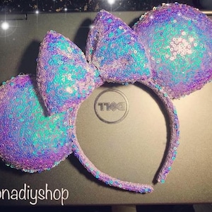May include: Purple and blue iridescent sequin Minnie Mouse ears with a large bow. The headband is white.
