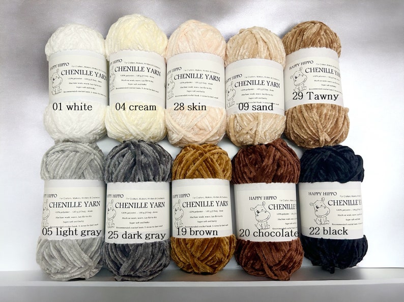 Op de afbeelding: Tien spoelen chenille garen in verschillende kleuren, waaronder wit, cr&egrave;me, huid, zand, bruin, lichtgrijs, donkergrijs, bruin, chocolade en zwart. Elke spoel heeft een label met de merknaam "Happy Hippo" en het kleur nummer en de kleur naam.