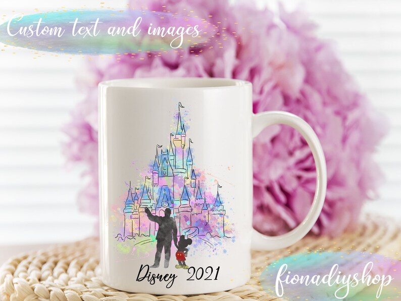 May include: White ceramic mug featuring a watercolor illustration of a castle with a person and Mickey Mouse. The mug has the text "Disney 2021" and "fionadiyshop" printed on it. The background includes a pink flower arrangement.