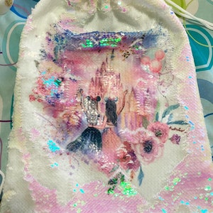 May include: White sequined drawstring bag featuring a colorful illustration of two figures in front of a castle, with pink and purple floral accents. The bag has a white drawstring closure and is covered in iridescent sequins.